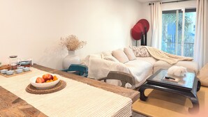 Un salon comprenant un canapé, une table basse, une table à manger avec un saladier de fruits, un vase de fleurs séchées et une fenêtre avec des rideaux.