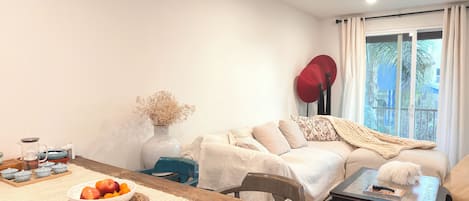 Un salon comprenant un canapé, une table basse, une table à manger avec un saladier de fruits, un vase de fleurs séchées et une fenêtre avec des rideaux.