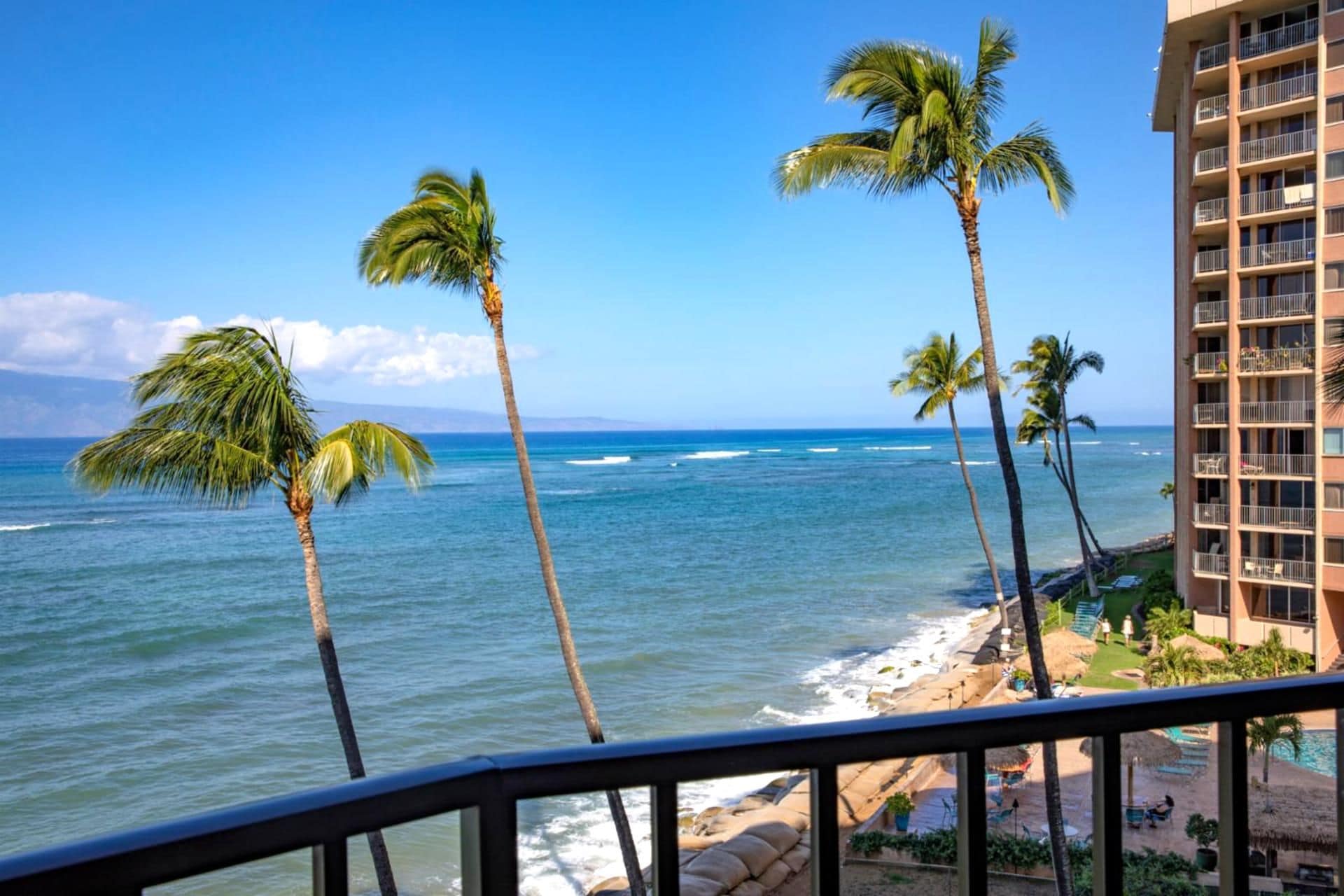 A beachfront view with palm trees, ocean waves, and a building with balconies.