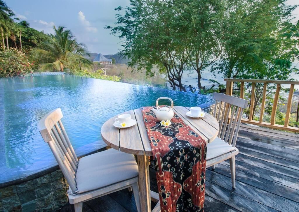 An infinity pool with a wooden deck, a table with a tablecloth, chairs, and a teapot.