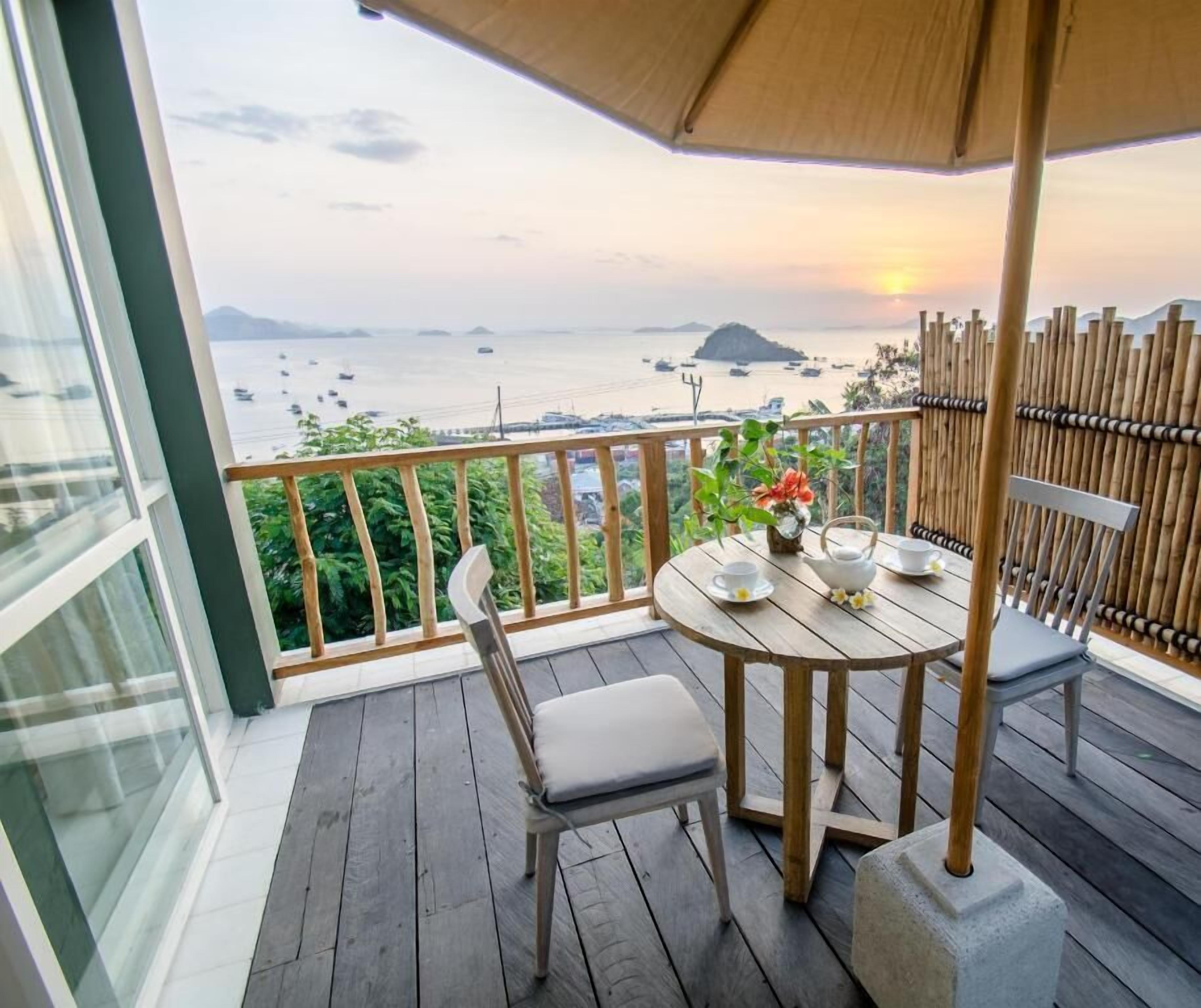A wooden deck with a table and chairs, a small table with a teapot and cups, and a view of the sea and boats.