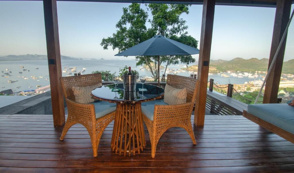 A wooden deck with wicker chairs and a table, overlooking a body of water with boats.