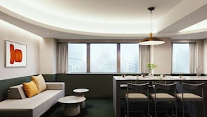 Una habitación de hotel moderna con distribución curva, un comedor con una mesa y sillas redondas, un sofá con cojines y un ventanal grande con vistas a la ciudad.