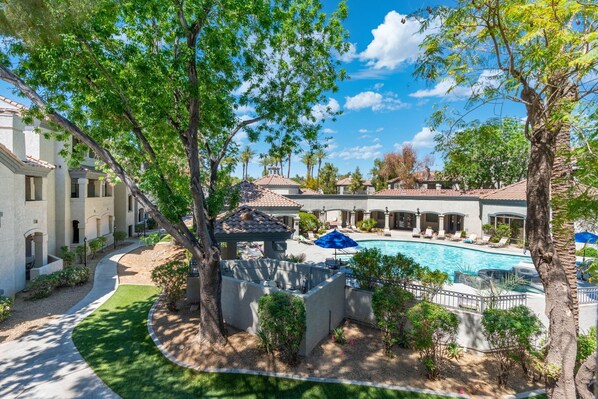A pool area with lounge chairs and umbrellas, surrounded by trees and a building with a tiled roof. - Spacious 2BR 2BA Condo Getaway for 6 (Scottsdale)