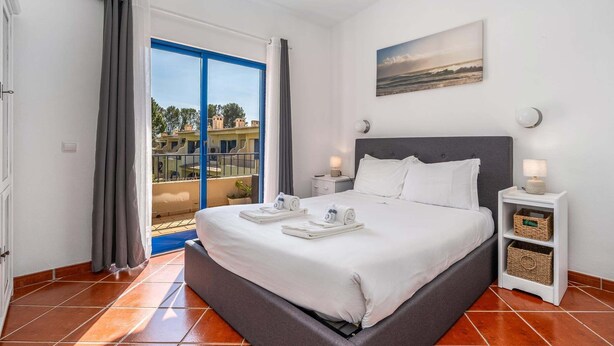 Un dormitorio con cama, mesita de noche, lámpara de pared, un cuadro y vistas de edificios a través de una ventana.