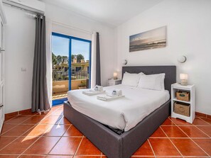 A bedroom with a bed, a nightstand, a wall-mounted lamp, a painting, and a view of buildings through a window. - Pool retreat at Clube da Bemposta (Alvor)