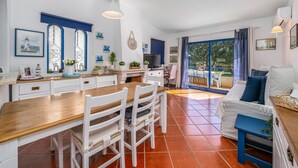 Una cocina moderna con una mesa de madera y sillas blancas, una zona de comedor con un sofá azul y un balcón con vistas a árboles.
