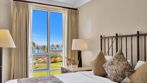 Quarto de hotel com uma janela grande, uma cama com roupa de cama floral, duas luminárias de cabeceira e uma mesa de cabeceira com um vaso de flores.