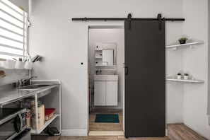 A kitchen with a microwave, sink, and storage shelves; a sliding door to a bathroom; and a small green plant on a shelf. - Miramar | Studio | Near T-mobile (San Juan)
