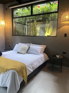 Un dormitorio con un ventanal grande, una cama con ropa de cama blanca, una mesita de noche pequeña y una manta amarilla.