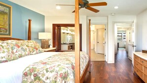 A bedroom with a four-poster bed, wooden flooring, a ceiling fan, and a hallway leading to a bathroom. - Halii Kai | Ocean View 7 BR, Sleeps 20 | Car Incl. w/ 6+ Nights | HLI ML-3872 by KBM (Waikoloa)