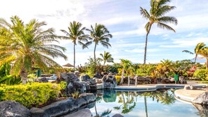 A pool area with lounge chairs, palm trees, and a waterfall. - Halii Kai | Ocean View 7 BR, Sleeps 20 | Car Incl. w/ 6+ Nights | HLI ML-3872 by KBM (Waikoloa)