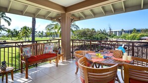A covered outdoor area with a wooden bench, a dining table with chairs, and a view of a golf course and buildings in the background. - Halii Kai | Ocean View 7 BR, Sleeps 20 | Car Incl. w/ 6+ Nights | HLI ML-3872 by KBM (Waikoloa)