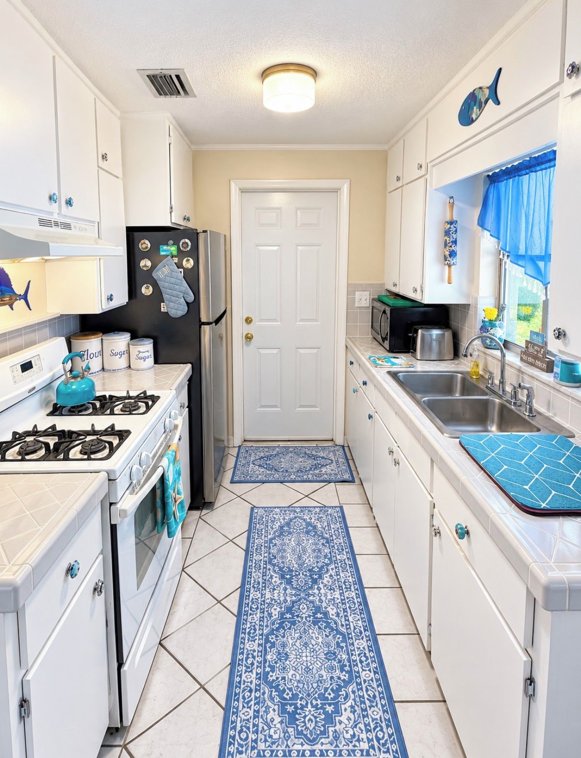 A compact kitchen with white cabinets, a stove, a refrigerator, and a microwave. There is a blue patterned rug on the floor.