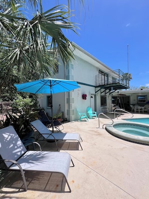 A pool area with lounge chairs, a blue umbrella, and a building with a balcony. - Tropical Riverside Oasis with Private Pool • Near drive-on beach & river access  (Edgewater)