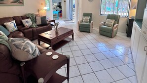 A living room with a sofa, coffee table, and armchairs, a ceiling fan, and a kitchen area in the background.