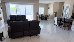 A spacious living area with a large dark-colored sofa, a dining table with chairs, a kitchen area with bar stools, and a sliding glass door leading to a balcony.
