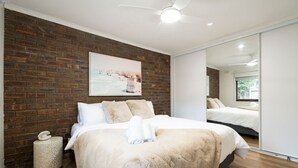 A modern bedroom with a brick wall, a ceiling fan, a large mirror, and a bed with white bedding. - Light filled 2 bedroom central retreat (Torquay)