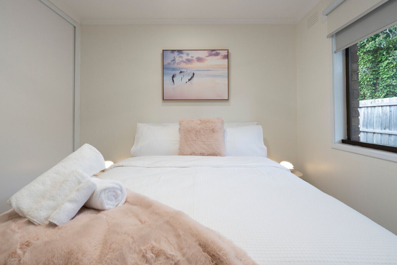 Un dormitorio con cama, almohadas, manta, un cuadro y una ventana.