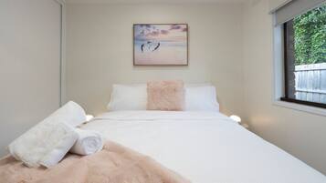Un dormitorio con cama, almohadas, manta, un cuadro y una ventana.
