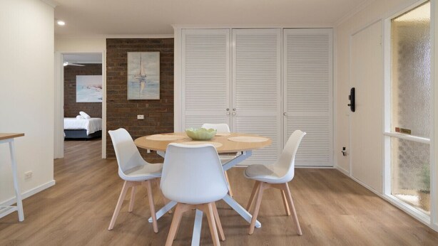 A dining area with a round table, white chairs, and a bowl. There is a brick wall with a painting, a bed in the background, and a door with louvered panels. - Light filled 2 bedroom central retreat (Torquay)