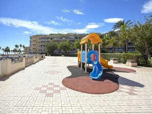 Un parc infantil amb tobogan i estructures per escalar, envoltat d'una zona pavimentada amb palmeres i un edifici al fons.