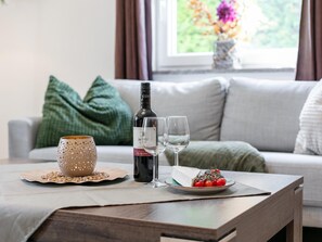 Une table en bois, une bouteille de vin, deux verres, une tasse à pois, une assiette de tomates cerises et un coussin vert sur un canapé.