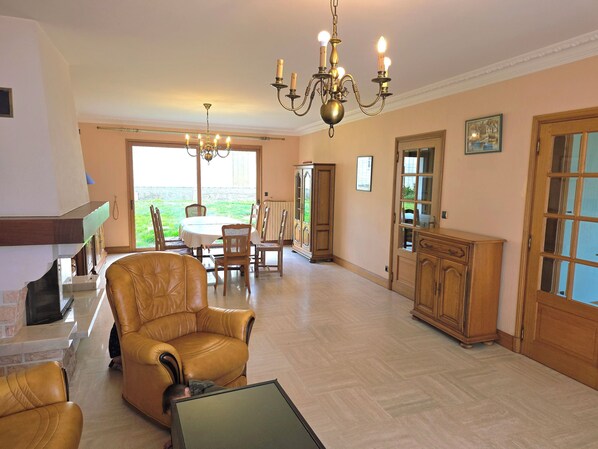 A spacious living room with a fireplace, a dining area with a table and chairs, a chandelier, and a view through a glass door. - Bretagne large seaside house (Moëlan-sur-Mer)