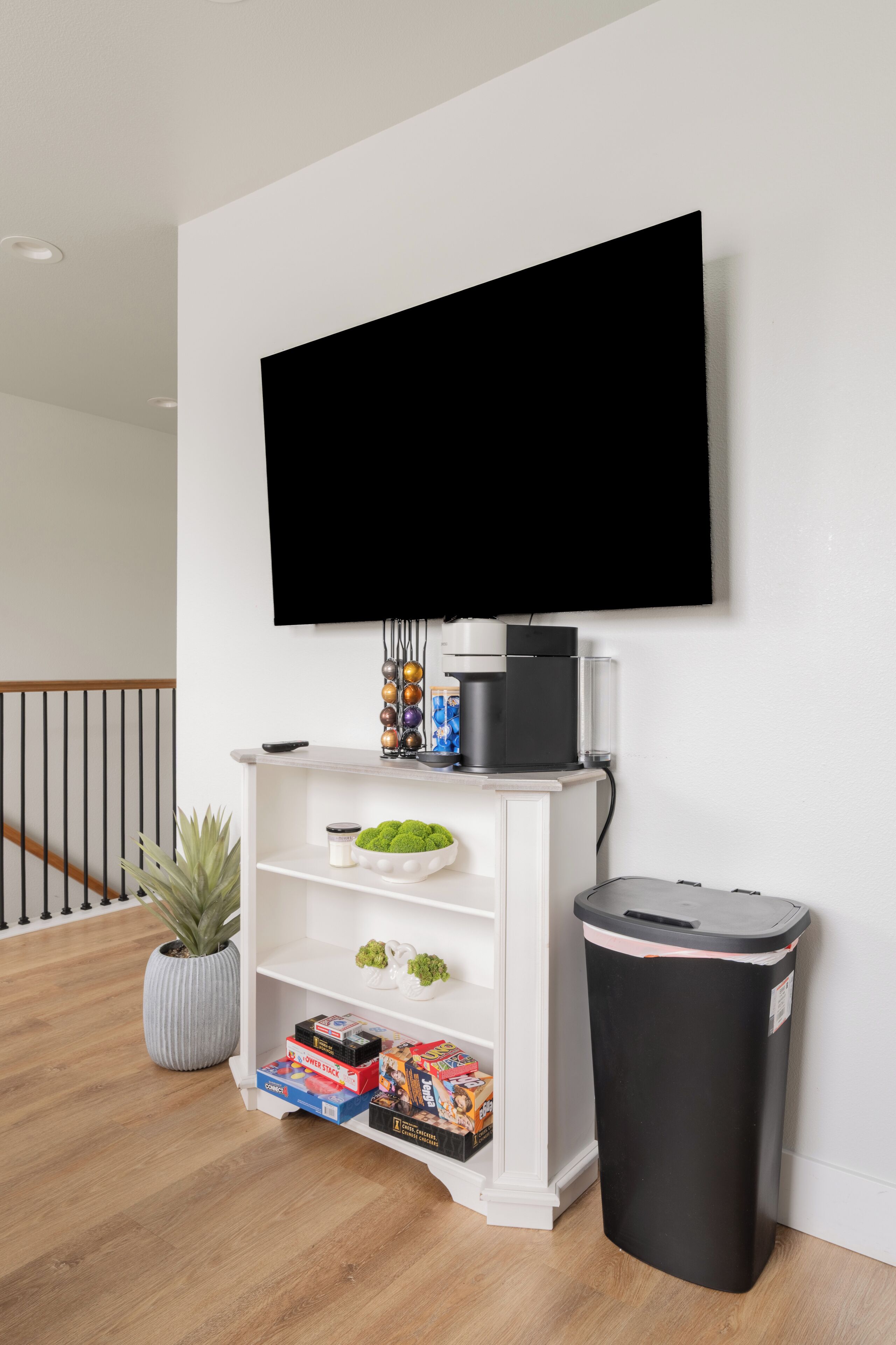 A living room with a mounted TV, a coffee maker, a trash can, and a shelf with decorative items and board games.