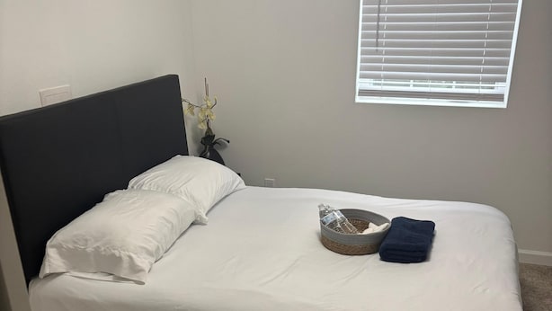 A single bed with white bedding, a dark headboard, a window with blinds, and a small vase with flowers. - Saffire Villa  (Tacoma)