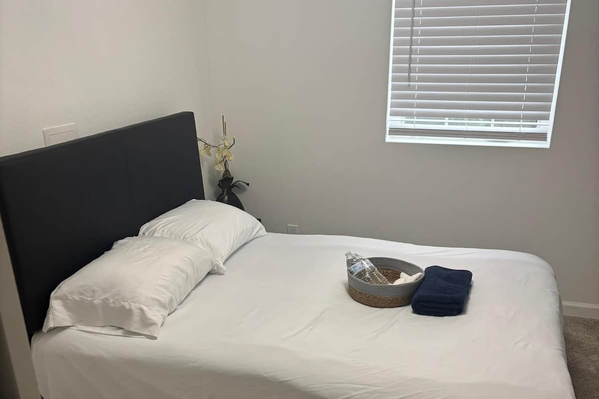A single bed with white bedding, a dark headboard, a window with blinds, and a small vase with flowers.