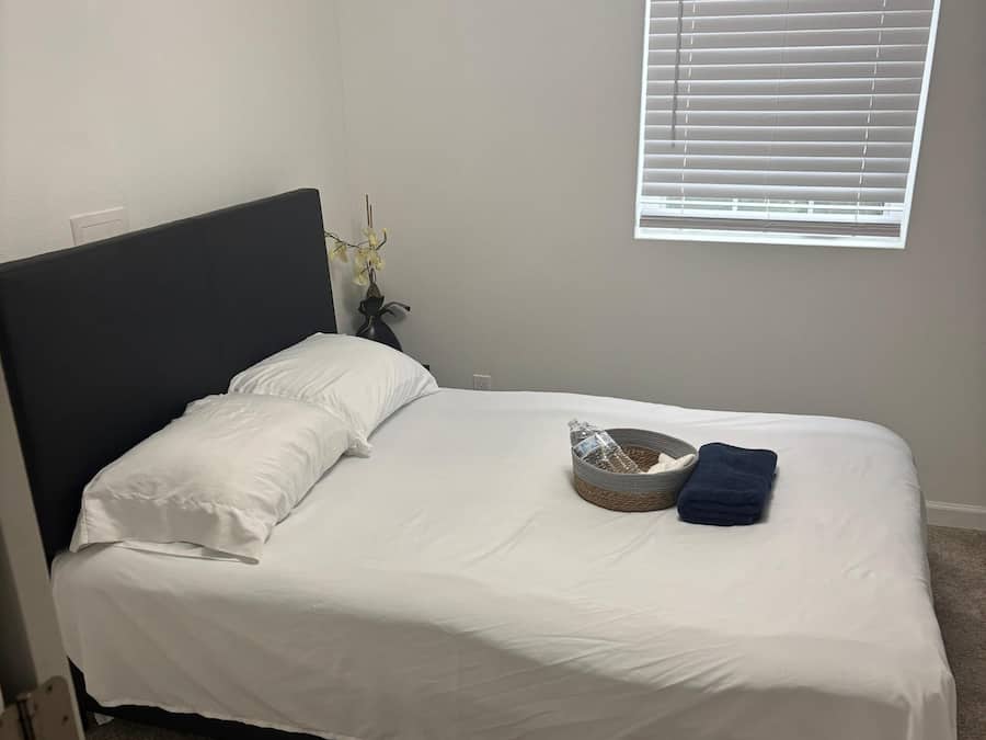 A single bed with white bedding, a dark headboard, a window with blinds, and a small vase with flowers.