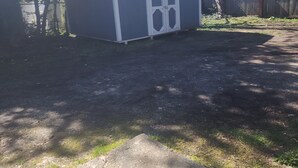 A small, single-story shed with a white door and windows, surrounded by a grassy yard and a wooden fence. - Saffire Villa  (Tacoma)