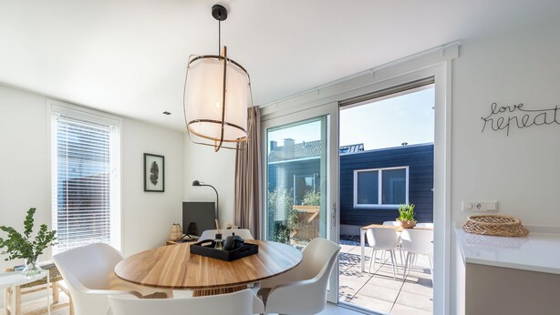 Een moderne eetruimte met een ronde houten tafel, witte stoelen, een hanglamp en uitzicht op buiten.