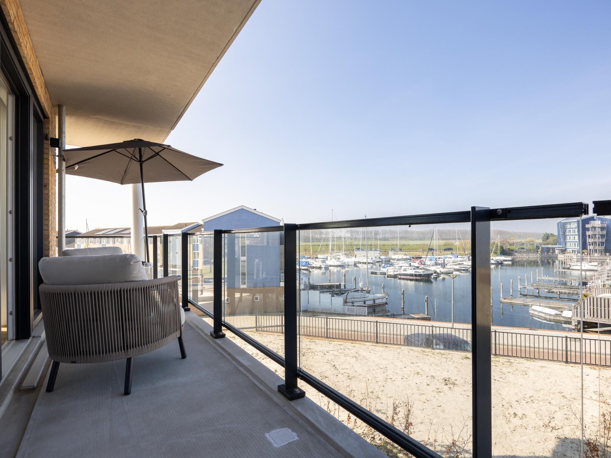 A balcony with a chair and umbrella, overlooking a marina with boats and buildings.