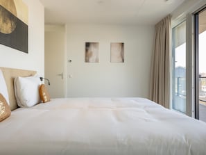 Une chambre d’hôtel avec un grand lit, une porte, deux  œuvres d’art mural et une vue par une grande fenêtre.