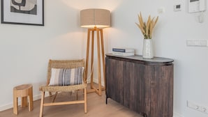 A modern room with a standing lamp, a wicker chair, a small stool, a side table with a vase, and a framed abstract artwork. - Luxury penthouse with terrace and harbour views (Kortgene)