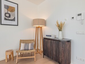 Ein modernes Zimmer mit Stehlampe, Korbsessel, kleines Hocker, Beistelltisch mit Vase und einem gerahmten abstrakten Kunstwerk.