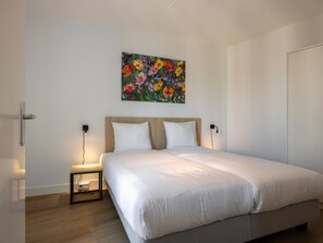 A modern bedroom with a large bed, a wooden headboard, a bedside table with a lamp, and a colorful floral painting on the wall. - Harbour apartment with beautiful views (Kortgene)