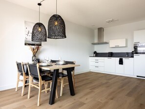 A modern kitchen with white cabinets, a black countertop, and a dining area with a wooden table and chairs. - Stylish harbour apartment with balcony & view (Kortgene)