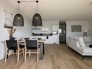 A modern kitchen with white cabinets, a dining area with a wooden table and chairs, and a living room with a sofa. - Stylish harbour apartment with balcony & view (Kortgene)
