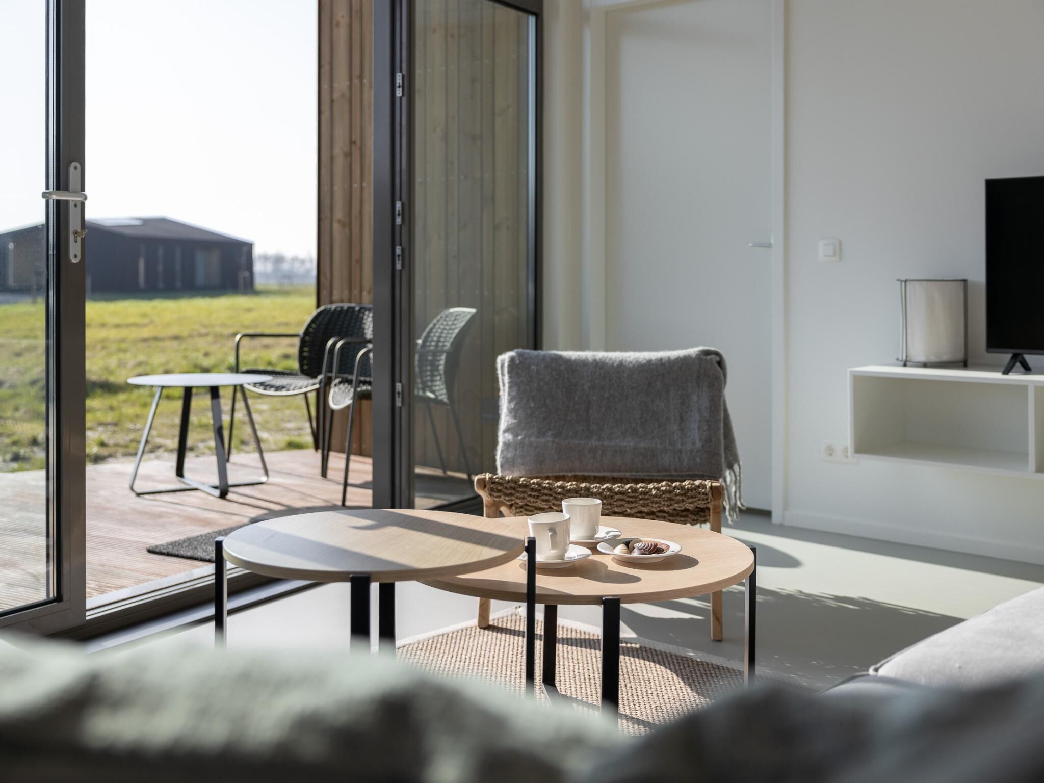 Een moderne woonkamer met een bank, een ronde tafel en een stoel. Er staat een televisie op een dressoir en een raam met uitzicht op het landschap.