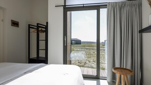 Un dormitorio con cama, un taburete de madera, una puerta corrediza de vidrio y vista a un edificio exterior.
