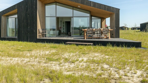 Ein modernes Haus mit einem großen Glasfenster, einer Holzterrasse und einer grünen Landschaft.