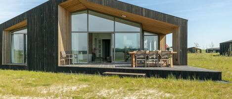 Una casa moderna con un amplio ventanal de vidrio, una terraza de madera y un paisaje cubierto de césped.