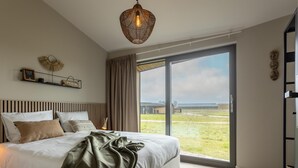 A modern bedroom with a large bed, a hanging light fixture, and a view of the countryside through a sliding glass door. - Nature house with sauna by nature reserve (Wissenkerke)