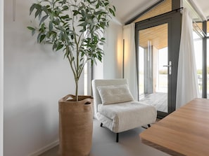 Una habitación con una planta grande en una cesta tejida, una butaca tapizada de blanco y una mesa de madera.