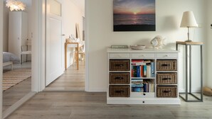 Una habitació amb sòl de fusta, una taula de trucat blanca amb cistos de vimbles, un quadre amb una imatge de sol ixent i una làmpada sobre una taula auxiliar.