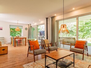 A spacious living area with a wooden dining table, two sofas with orange cushions, a coffee table, and large windows offering a view of greenery. - Nice home close to the beach (100 meters) (Kamperland)