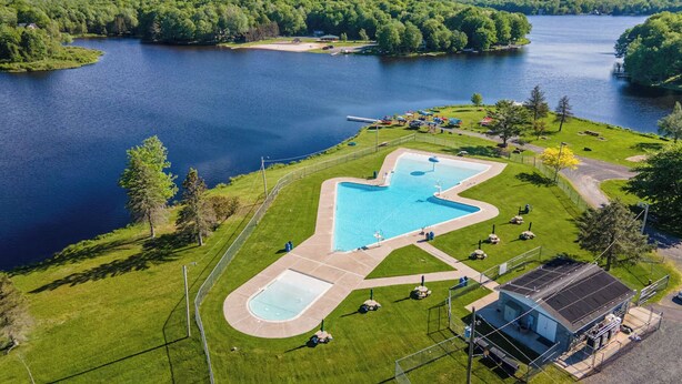 Oversigtsbillede af en ejendom ved en sø med swimmingpool, en mindre bygning og grøn bevoksning omkring.